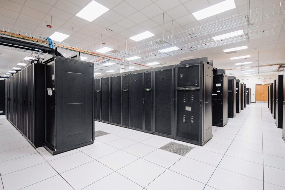 Data hall inside a data center, with rows of server cabinets lining a cold aisle under overhead cable trays.